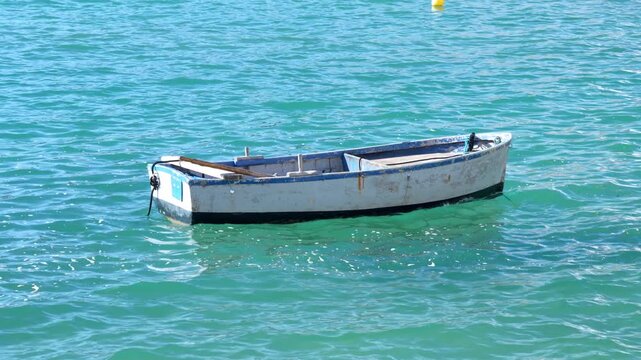 An empty wooden rowboat with peeling white and blue paint bobs gently on rippling turquoise water in a sunny bay, moored with a rope beside a small yellow buoy, a peaceful coastal scene.