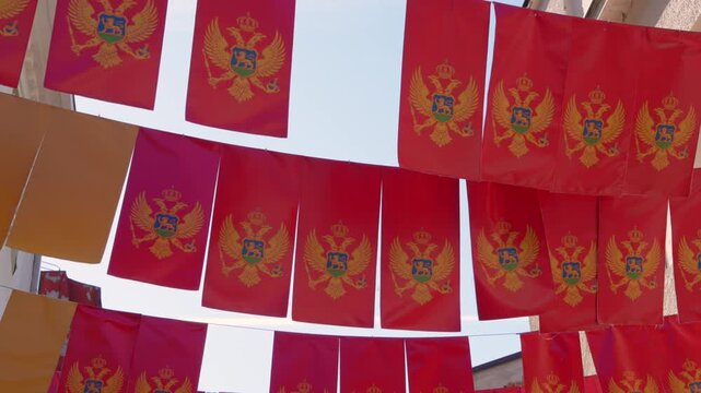 Rows of red Montenegrin flags with a golden double headed eagle hang on strings above a street and sway gently against a bright sky, a concept of national pride and independence.