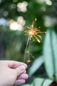 Hand holding lit sparkler emitting bright spark firework light glow during festive outdoor celebration in green garden with bokeh background creates joyful hopeful mood