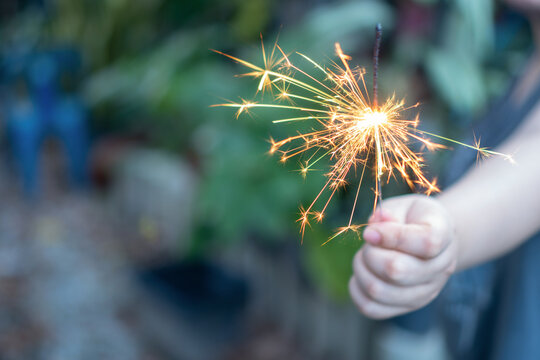 Child hand holding burning sparkler firework with bright light and glowing spark during outdoor celebration, showing happy emotion during night event in garden party setting