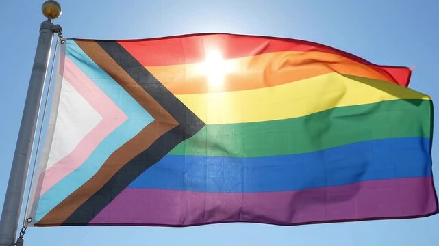 Progress Pride Flag waving in the wind against blue sky with bright sun flare, LGBTQ+ inclusion and diversity concept.