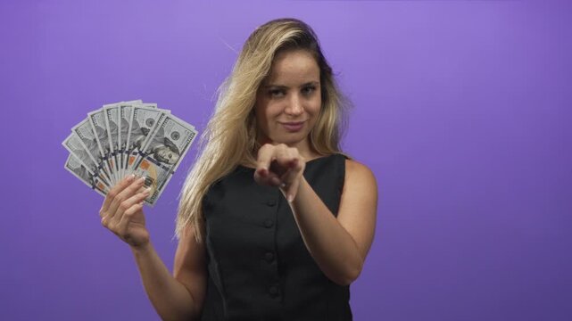 Young blonde woman holding a fan of us dollar cash and pointing finger toward camera in purple studio; confidence wealth success.