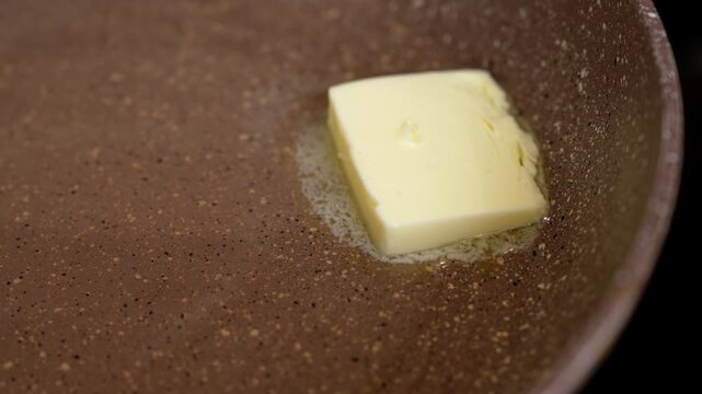 A square piece of butter softens and melts on the speckled surface of a brown nonstick frying pan in a home kitchen, while a knife slices off more, a cooking preparation step.