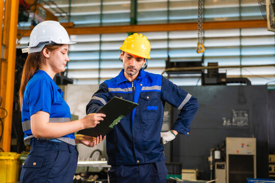 Factory workers reviewing production report on clipboard in manufacturing plant, industrial engineers in safety gear discussing quality control data in workshop