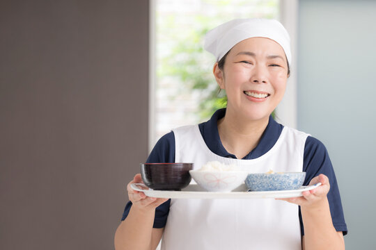 高齢者の自宅で三角巾をつけ食事を用意する笑顔の女性スタッフ、家事代行、料理代行、訪問介護、食事支援、配膳イメージカメラ目線笑顔