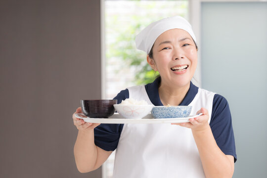 高齢者の自宅で三角巾をつけ食事を用意する笑顔の女性スタッフ、家事代行、料理代行、訪問介護、食事支援、配膳イメージカメラ目線笑顔