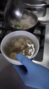 Serving homemade meatball soup, pouring hot broth into a white bowl in a kitchen.