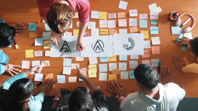 Top down aerial view of skilled designer hold logo while discuss with team. Aerial business group working together sharing idea about logo graphic design by using sticky notes and tablet. Symposium.