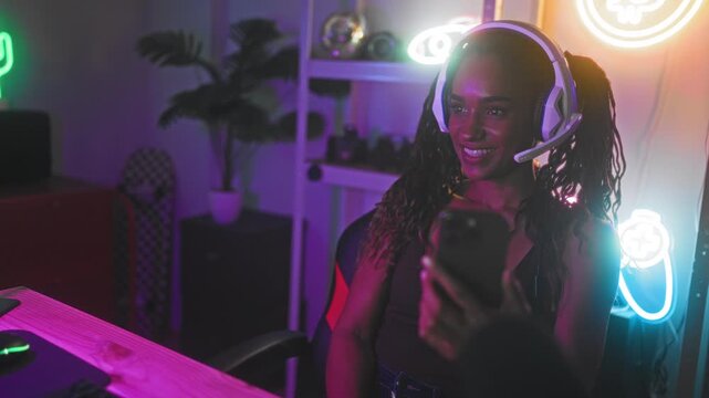 Woman wearing gaming headset holds smartphone, smiles and waves hand toward camera in neon streamer studio with shelves and skateboard; friendly connection joy.