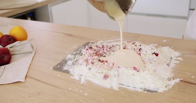 Berry pastry preparation. Baking cake. Female chef pouring liquid batter from bowl in flour crumbs cranberry pile on wooden table.