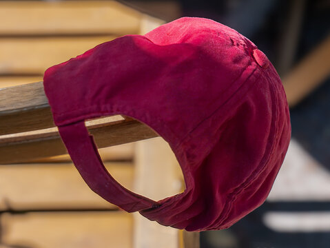 Crimson baseball cap hanging on the arm of a wooden chair, rear view with adjustable strap in sunlight