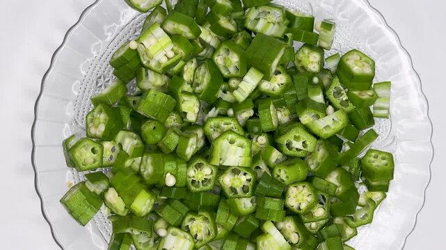 Rotating top view of fresh chopped okra pieces on transparent plate over white background.