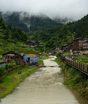 Village path and creek lined with wooden houses under misty mountains, near Xijiang, Guizhou, China
