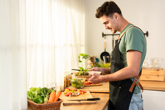 Latino man wearing kitchen apron, preparing fresh salad in kitchen.