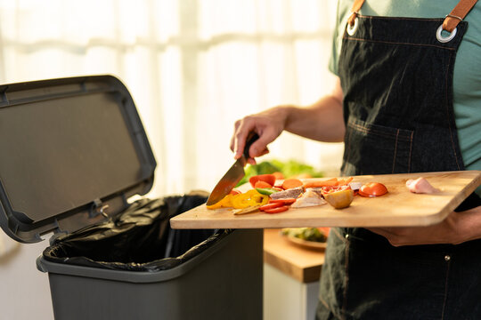 Close up of man in kitchen apron throwing away fresh food scraps into trash bin.