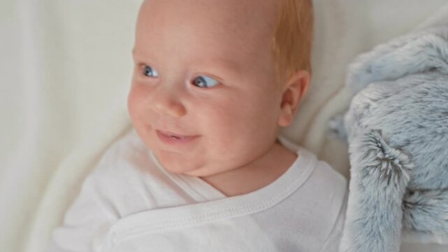 Baby lying on bed in onesie with open mouth and plush toy, smiling and gazing in studio; gentle joy.