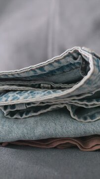Close up view of folded clothing items including brown fabric and blue denim jeans on flat surface. hand places additional garments gradually building stack