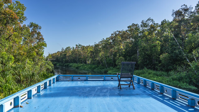An Indonesian boat, a klotok, is floating along the river in the jungle. There is a wooden chair on the upper deck. The banks are overgrown with green plants &mdash; grass, trees. The blue sky. Borneo.

 