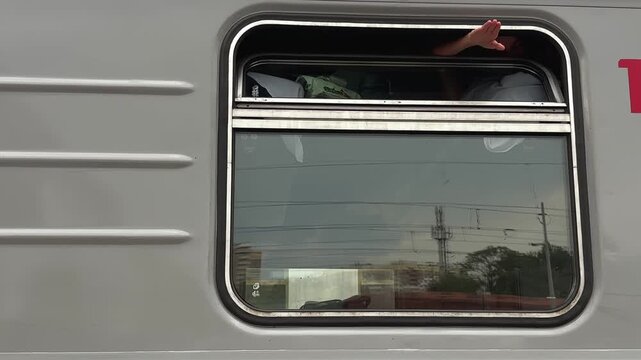 A teenage girl is lying on the upper bunk in a train compartment and waving her hand through the open window. A journey, a train trip. A view from the platform.