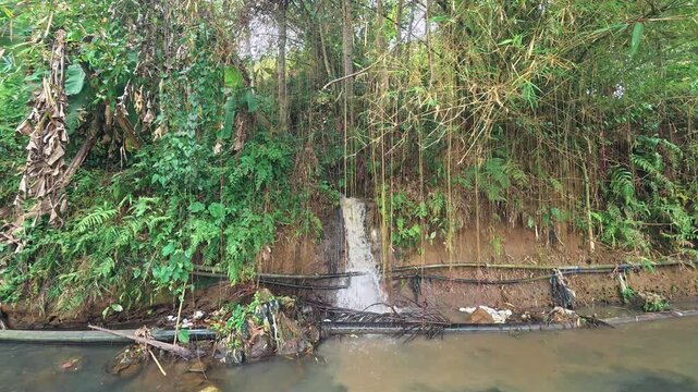 Cinematic footage of a small water runoff flowing down a dirt riverbank covered in lush green vegetation and bamboo into a murky stream
