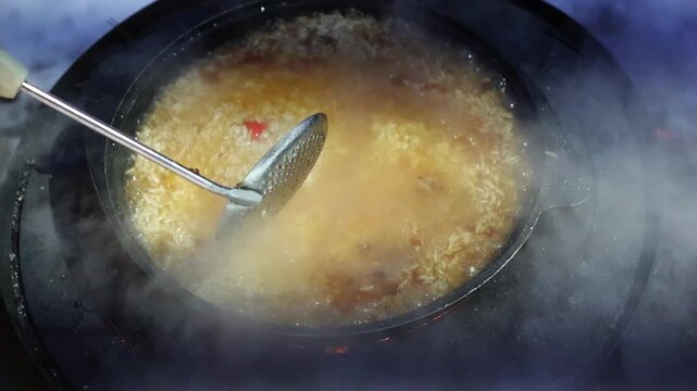 Close-up of plov being stirred in a steaming pan during outdoor winter cooking. Rice, broth and spices simmer over a hot stove with fire glowing beside the snowy cooking area.