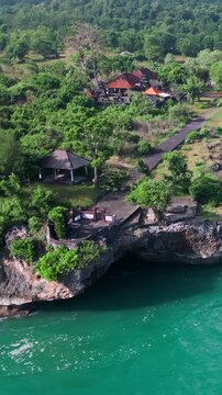 Aerial bali cliff coastline ocean view lush tropical vegetation crowns jagged limestone cliffs with hidden sea cave, emerald water, gentle waves lapping rocky shore, Jimbaran, Bali, Vertical video
