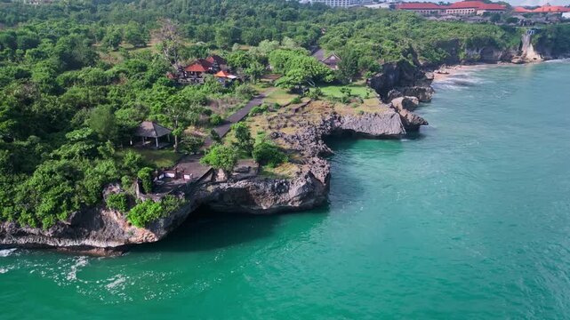 Aerial bali cliff coastline ocean view lush tropical vegetation crowns jagged limestone cliffs with hidden sea cave, emerald water, gentle waves lapping rocky shore, Jimbaran, Bali, Vertical video