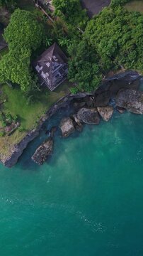 Aerial view bali temple cliff overlooking turquoise ocean, drone captures cliffside shrine surrounded by lush greenery, rocky cove and emerald lagoon, waves lapping against stone, vertical video