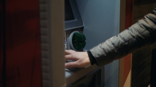 Close up shot showing hand inserting credit card into ATM machine slot and typing PIN code upon keypad for cash withdrawal or banking transaction