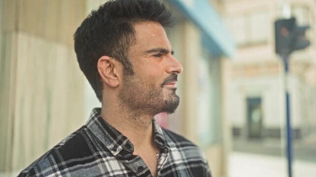 Man profile with beard and visible ear looking right in a plaid shirt on a city street beside a building facade; quiet contemplation.