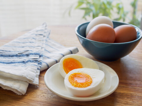 Soft boiled egg cut in half on a white plate with blue striped linen cloth and eggs in a bowl on a wooden table by the window