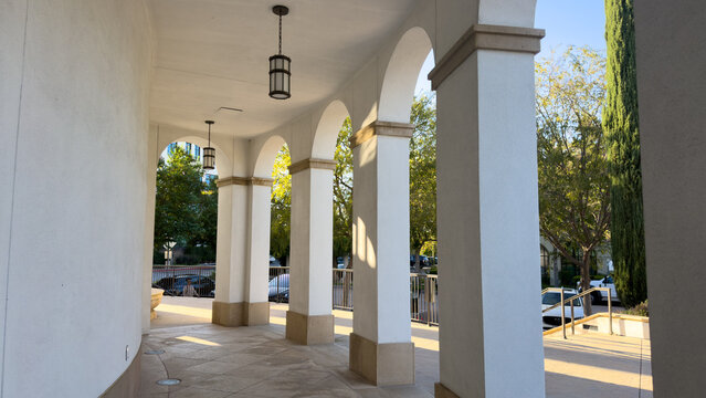 an elegant outdoor covered walkway features white stucco arches square pillars and hanging lantern lights overlooking trees and parked cars