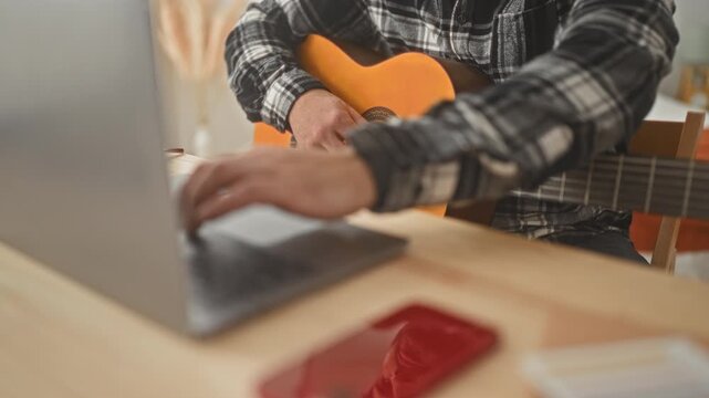 Man hispanic playing classical guitar with fingers on fretboard and right hand strumming beside a laptop on a desk in a studio apartment with plants and lamp; concentration.