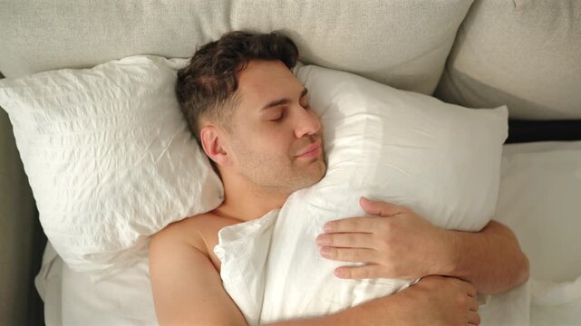 A shirtless man hugs a white pillow in bed, opens his eyes with a content smile, then closes them again as camera pushes in from overhead. Warm morning sun on seersucker pillows and beige headboard.