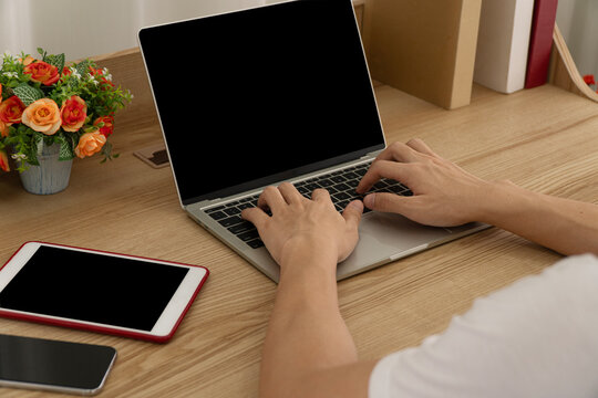 Over the shoulder close up view of man hands typing on laptop computer with blank black screen on wooden desk. Tablet, smartphone and home office workspace mockup presentation concept.