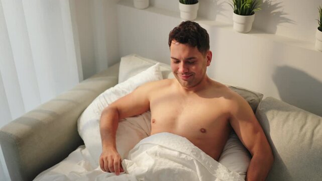 Shirtless young man wakes up smiling under white duvet on a gray sofa bed, then picks up a teal smartphone to scroll messages. Sunlight through sheer curtains, potted plants on shelf. Happy morning.
