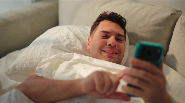 Smiling man lies in bed under a white duvet and scrolls a teal phone with his finger. Happy guy texting and chatting online on a beige sofa bed with seersucker pillow. Cozy relaxed home lifestyle.