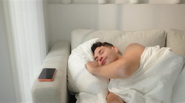 Sleepy man asleep on sofa bed under white duvet reaches for orange smartphone on armrest. Tired man squints at phone screen with eyes half closed. Sunny morning wake up routine in bright bedroom.