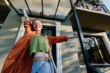Happy young woman stretching arms with joy on the porch of a house at sunrise. Wearing an orange...