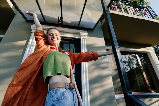 Happy young woman stretching arms with joy on the porch of a house at sunrise. Wearing an orange coat and green top. Concept of freedom, morning energy, and new day.