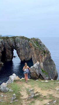 woman tourist standing on cliff looking at natural rock arch on coast in spain, Asturias, vertical video
