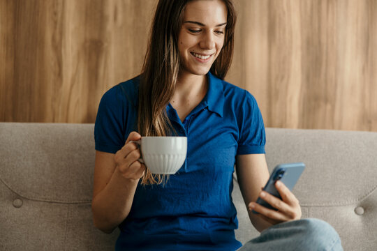 Woman smiling using smartphone and drinking coffee at home