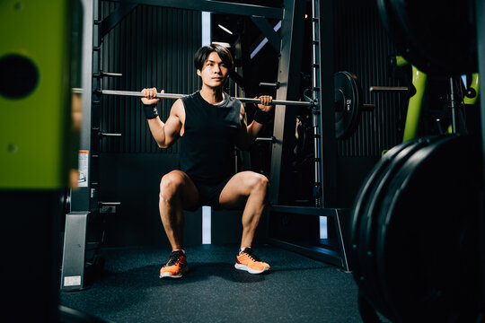 Asian muscular man guy performing a squat exercise legs heavy weights with a barbell in gym interior, male doing lifting barbell work out at fitness gym, sport workout healthy lifestyle
