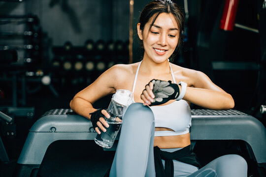 Portrait of beautiful Asian young woman sitting resting holding a bottle of water and check time on smart watch in fitness gym, female workout sport motivation, healthy lifestyle