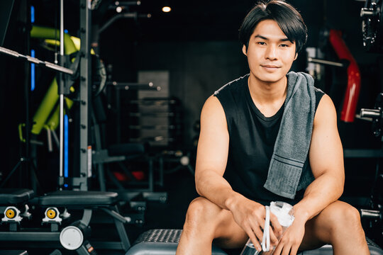 Portrait of Asian young man trained body in black sportswear sitting for rest holding water or protine in fitness gym, Weight training, healthy lifestyle
