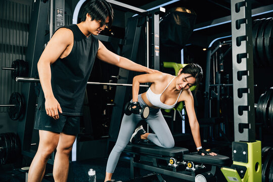 Trainer man teaching woman lifting dumbbell in fitness gym, Happy Asian coach training young female help with a difficult exercise at personal gym, Weight training