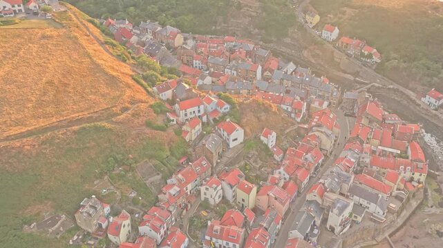 Staithes village and Staithes Beck extend between steep coastal ravines and farmland, with dense red roofed cottages and winding roads, drone forward flyover with descent