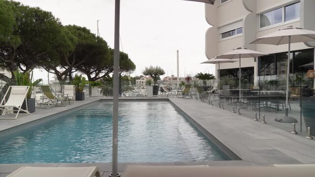 A lengthwise view shows an outdoor swimming pool beside a modern resort building, with sun loungers and trees on one side and a dining terrace on the other.
