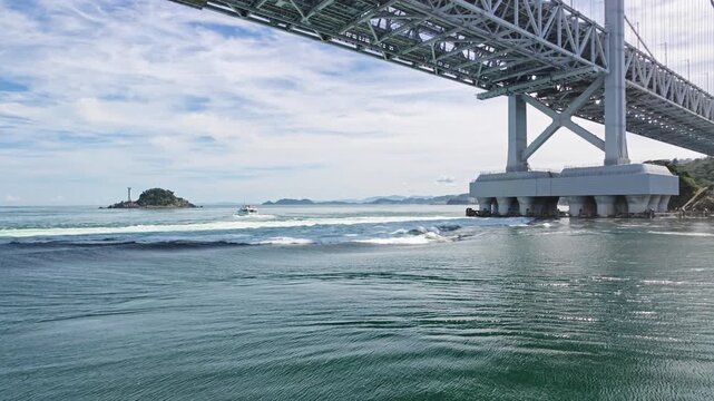 Low-angle view looking up at massive bridge steel truss framework and piers with swirling tidal waves and a distant lighthouse islet below.
