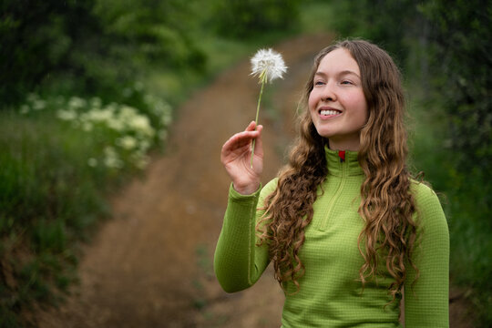 Smiling curly haired woman holding a dandelion and admiring the flower while standing on a lush green trail surrounded by nature.